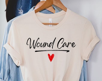 Puede incluir: Una camiseta beige con el texto "Wound Care" en escritura negra con un corazón rojo debajo. La camiseta está colgada en una percha de madera.