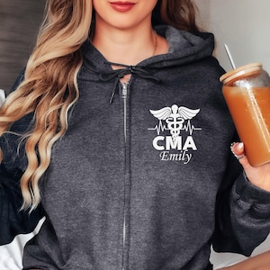 Puede incluir: Una sudadera con capucha gris oscuro con un símbolo de caduceo médico blanco, una línea de latido del corazón y el texto "CMA Emily".