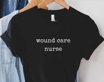Puede incluir: Camiseta negra con texto blanco que dice "wound care nurse" colgada de una percha de madera.