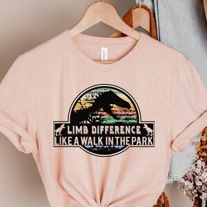Peut inclure: T-shirt rose clair avec un motif de dinosaure et le texte "LIMB DIFFERENCE LIKE A WALK IN THE PARK". Le motif est dans un cadre circulaire avec un fond de coucher de soleil coloré.