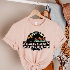 May include: A light pink t-shirt with a graphic featuring a dinosaur silhouette within a circular design. The text reads "BLEEDING DISORDER LIKE A WALK IN THE PARK". The shirt is hanging on a wooden hanger.