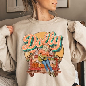 May include: Beige sweatshirt featuring a graphic of a woman playing a guitar in a truck bed filled with pumpkins. The word "Dolly" is written in large, retro-style lettering above the image. The design has a vintage, country aesthetic.