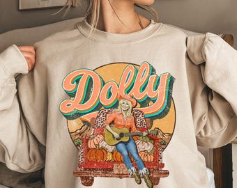 May include: Beige sweatshirt featuring a graphic of a woman playing a guitar in a truck bed filled with pumpkins. The word "Dolly" is written in large, retro-style lettering above the image. The design has a vintage, country aesthetic.