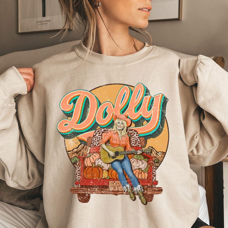 Ivory T-shirt with a retro distressed Dolly design featuring her in an orange cowgirl shirt, blue jeans, boots, and hat, sitting on a truck bed decorated with pumpkins and cow print blankets. Large vintage “Dolly” lettering above with teal and orange retro style.