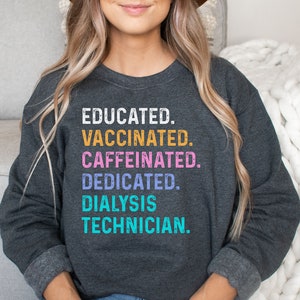 May include: A dark grey sweatshirt with the text "EDUCATED. VACCINATED. CAFFEINATED. DEDICATED. DIALYSIS TECHNICIAN." printed in colourful letters.