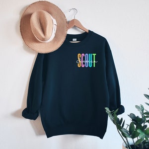 May include: A navy blue sweatshirt with the word "SCOUT" in colorful vertical letters and the name "Jessica" in cursive. A straw hat hangs on the wall next to the sweatshirt.