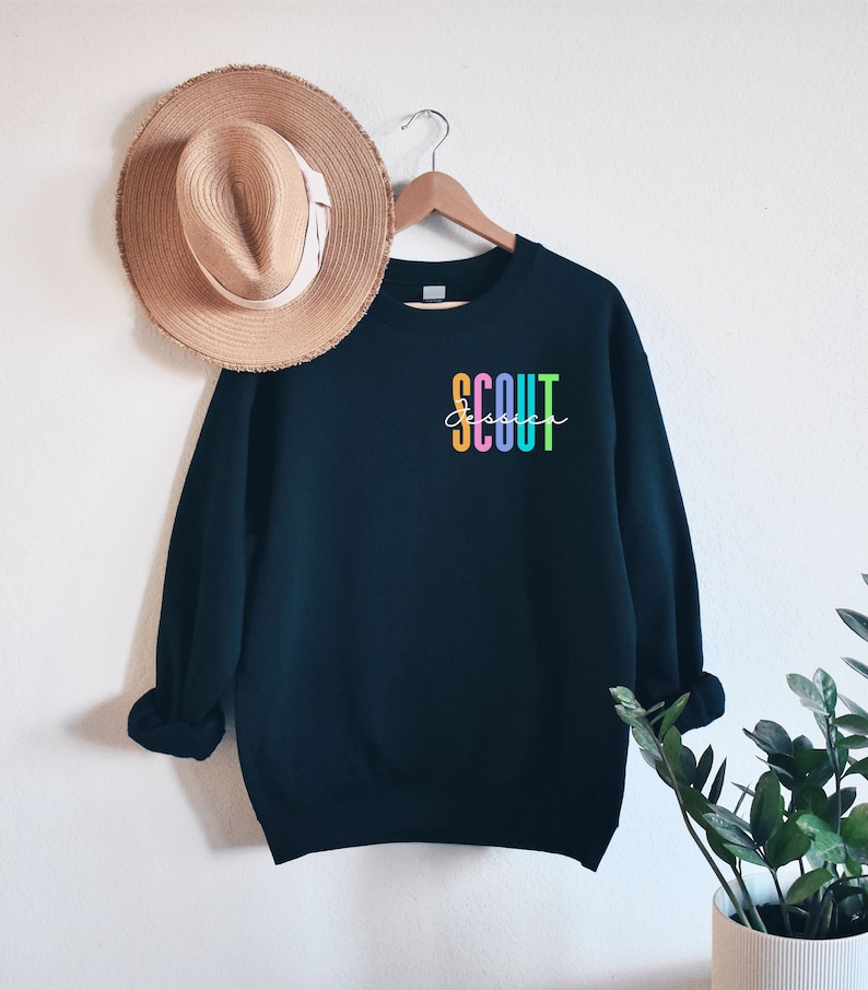 May include: A navy blue sweatshirt with the word "SCOUT" in colorful vertical letters and the name "Jessica" in cursive. A straw hat hangs on the wall next to the sweatshirt.