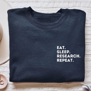 May include: A black sweatshirt with white text that reads "EAT. SLEEP. RESEARCH. REPEAT."