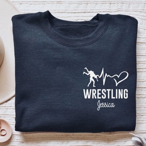 Könnte beinhalten: Marineblaues Sweatshirt mit weißer Grafik von zwei Ringern, einer Herzschlaglinie und einem Herz. Das Wort "WRESTLING" steht darunter, mit dem Namen "Jessica" darunter. Ein cremefarbener Hut und Schmuck sind im Hintergrund.
