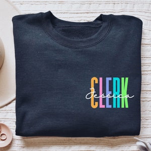 Puede incluir: Una sudadera negra con un gráfico de "CLERK" colorido y el nombre "Jessica" escrito en cursiva blanca.