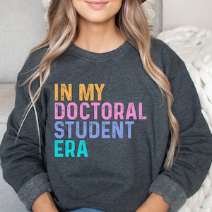 May include: A dark gray sweatshirt with the text "IN MY DOCTORAL STUDENT ERA" in colorful, distressed lettering.