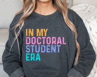 Może przedstawiać: Szara bluza z nadrukiem "IN MY DOCTORAL STUDENT ERA" w kolorowych, postarzonych literach.