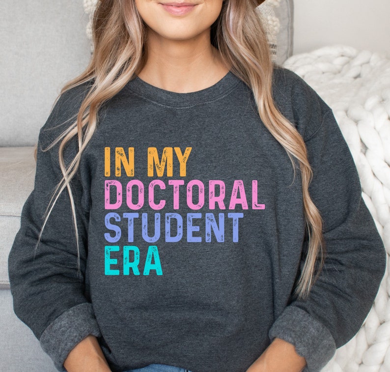 May include: A dark gray sweatshirt with the text "IN MY DOCTORAL STUDENT ERA" in colorful, distressed lettering.