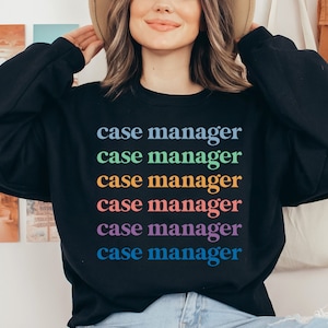 Op de afbeelding: Zwarte sweatshirt met de tekst "case manager" in regenboogkleuren. De tekst is verticaal gestapeld, waarbij elk woord een andere kleur heeft. De sweatshirt heeft een ronde hals.
