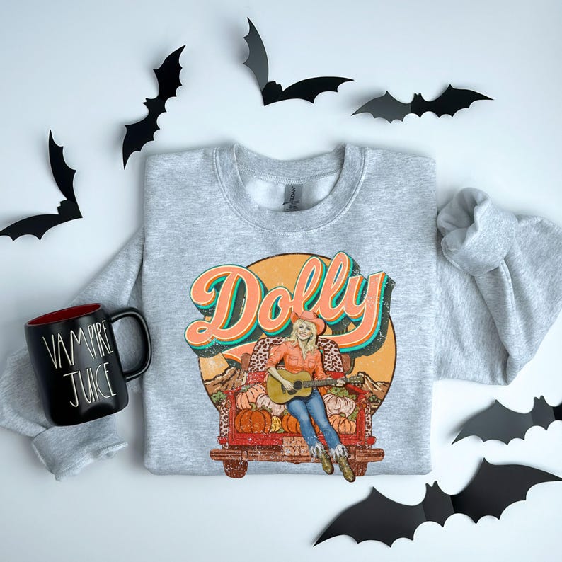 May include: A heather gray sweatshirt featuring a retro graphic of a woman playing a guitar in a truck filled with pumpkins, with the name "Dolly" in orange and teal lettering. A black mug with "VAMPIRE JUICE" text is on the left. Black bat cutouts are scattered around.