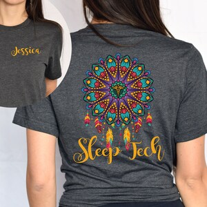 Può includere: T-shirt grigio scuro con il nome "Jessica" in oro sul davanti. Il retro presenta un colorato design mandala con le parole "Sleep Tech" in oro sottostanti.