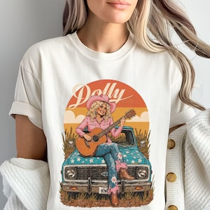 May include: Cream-colored t-shirt featuring a graphic of a woman in a pink shirt and hat playing a guitar, sitting on a blue truck. The design includes the name "Dolly" in a script font, set against a sunset backdrop.