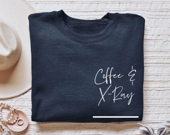 May include: A navy blue sweatshirt with the words "Coffee & X-Ray" in white script. A white hat, jewelry, and a ring are arranged around the sweatshirt on a textured surface.