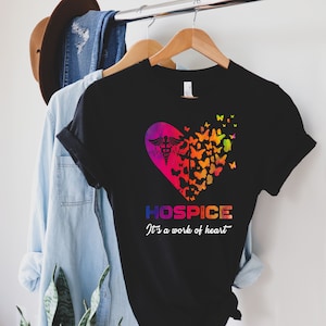May include: Black t-shirt with a rainbow heart design featuring a medical symbol and butterflies. The text "Hospice It's a work of heart" is printed below the heart.