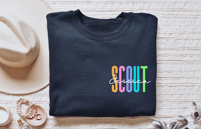 May include: Navy blue sweatshirt with the word "SCOUT" in colorful block letters and the name "Jessica" in cursive. A white hat and jewelry are visible in the background.