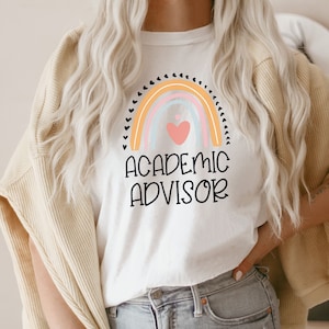 May include: White t-shirt with a rainbow design and the words "ACADEMIC ADVISOR" in black. The rainbow features pastel colors and a heart. The shirt is a casual style.