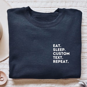 May include: A black sweatshirt with white text that reads "EAT. SLEEP. CUSTOM TEXT. REPEAT."