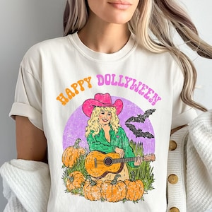 Puede incluir: Camiseta color crema con un gráfico que presenta a una mujer con un sombrero de vaquero rosa tocando la guitarra, rodeada de calabazas y murciélagos. El texto "HAPPY DOLLYWEEN" está arqueado en la parte superior en naranja y rosa.