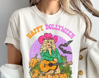 May include: Cream-colored t-shirt with a graphic featuring a woman in a pink cowboy hat playing a guitar, surrounded by pumpkins and bats. The text "HAPPY DOLLYWEEN" arches across the top in orange and pink.