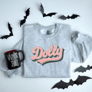 May include: A light gray sweatshirt with the name "Dolly" in a retro font, featuring pink and green accents. A black mug with "Vampire Juice" text and black bat decorations are also visible.