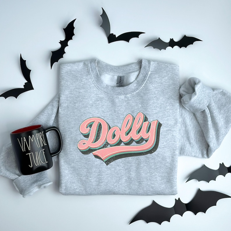 May include: A light gray sweatshirt with the name "Dolly" in a retro font, featuring pink and green accents. A black mug with "Vampire Juice" text and black bat decorations are also visible.