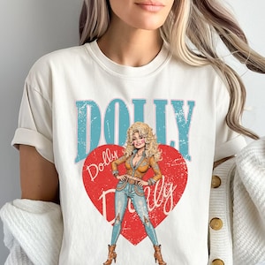 Puede incluir: Camiseta color crema con la palabra "DOLLY" en letras de bloque turquesas sobre un corazón rojo. Dentro del corazón hay una ilustración de una mujer con un atuendo de estilo occidental. La camiseta también tiene la palabra "Dolly" en cursiva.