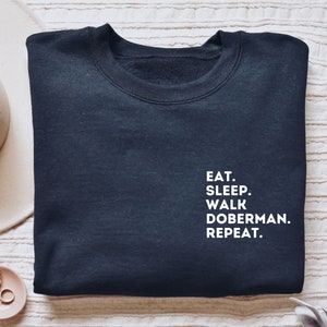 May include: A black sweatshirt with white text that reads "Eat. Sleep. Walk Doberman. Repeat."