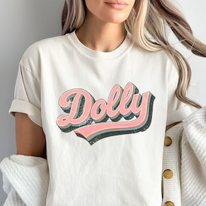 May include: Cream-coloured t-shirt with the name "Dolly" in a retro, layered design. The word "Dolly" is in a pink script with a teal outline and a black shadow, creating a vintage aesthetic. The shirt has short sleeves.