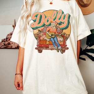 May include: Cream-colored t-shirt featuring a graphic of a woman playing a guitar in a truck bed filled with pumpkins. The word "Dolly" is written above the image in a retro font. The shirt has short sleeves and a relaxed fit.