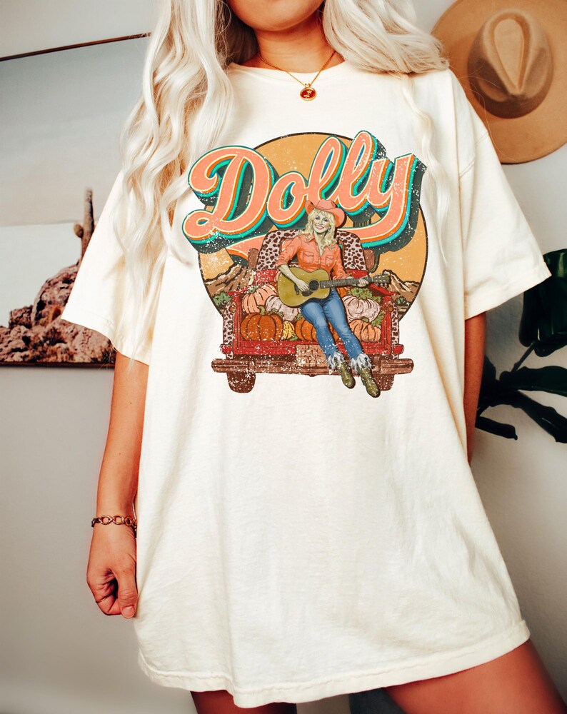 May include: Cream-colored t-shirt featuring a graphic of a woman playing a guitar in a truck bed filled with pumpkins. The word "Dolly" is written above the image in a retro font. The shirt has short sleeves and a relaxed fit.