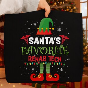May include: Black tote bag with the text "SANTA'S FAVORITE REHAB TECH" in green and white. The design includes an elf hat, medical symbols, snowflakes, and elf feet with red shoes. A string of colorful lights is also included.