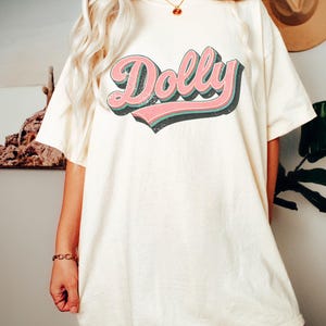 May include: Cream-colored t-shirt with the word "Dolly" in a retro font. The word is in pink with a black outline and a teal shadow. The shirt is a relaxed fit.