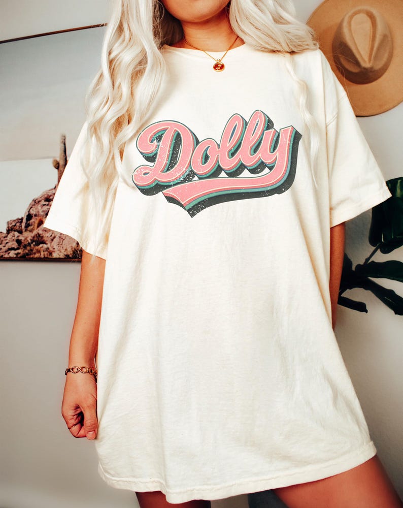 May include: Cream-colored t-shirt with the word "Dolly" in a retro font. The word is in pink with a black outline and a teal shadow. The shirt is a relaxed fit.