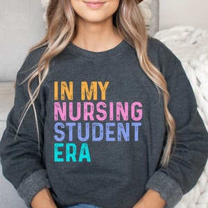 May include: Dark gray sweatshirt with the text "IN MY NURSING STUDENT ERA" in orange, pink, blue, and teal lettering. The sweatshirt has a crew neck and long sleeves with gray cuffs. The overall theme is nursing student apparel.