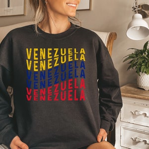 May include: Dark gray sweatshirt with the word "VENEZUELA" printed in yellow, blue, and red, representing the colors of the Venezuelan flag. The text is stacked vertically, creating a bold graphic design. The sweatshirt is a casual apparel item.