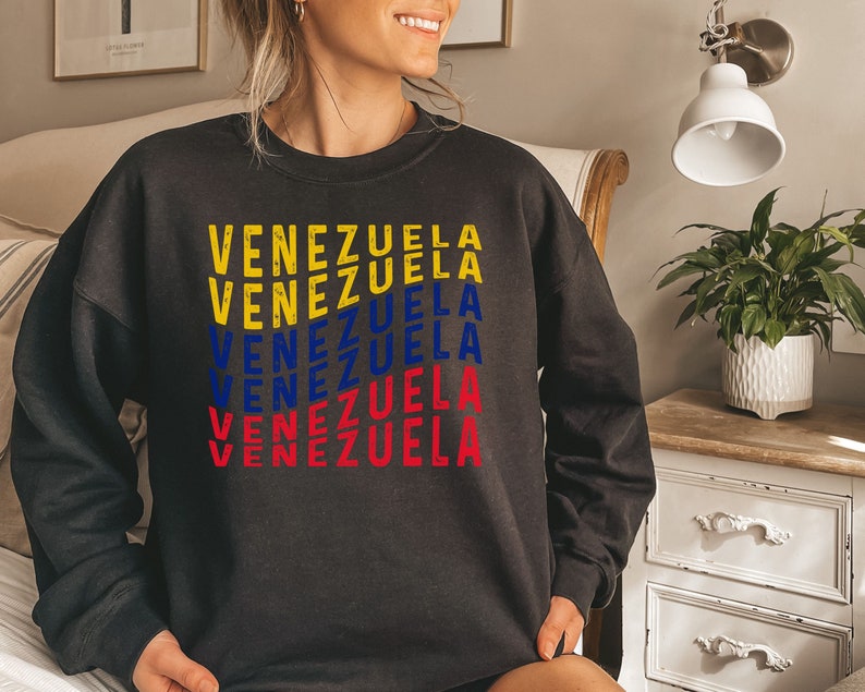May include: Dark gray sweatshirt with the word "VENEZUELA" printed in yellow, blue, and red, representing the colors of the Venezuelan flag. The text is stacked vertically, creating a bold graphic design. The sweatshirt is a casual apparel item.