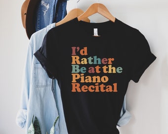 Puede incluir: Camiseta negra con el texto "I'd Rather Be at the Piano Recital" en un esquema de colores retro. La camiseta está colgada en una percha de madera con otras prendas en el fondo.
