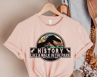 May include: A light pink t-shirt with a graphic featuring a dinosaur silhouette and the text "HISTORY LIKE A WALK IN THE PARK." The design includes a circular emblem with a sunset color scheme and a black rectangular banner.