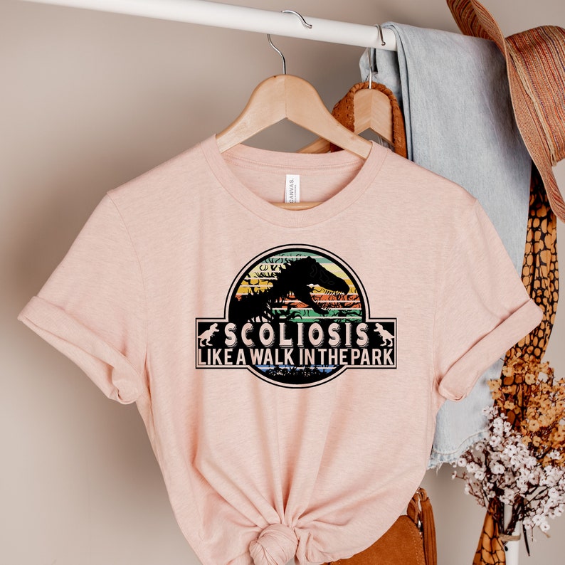 May include: A pink t-shirt with a graphic of a dinosaur skeleton in a Jurassic Park-style logo. The text "Scoliosis is like a walk in the park" is printed below the dinosaur.