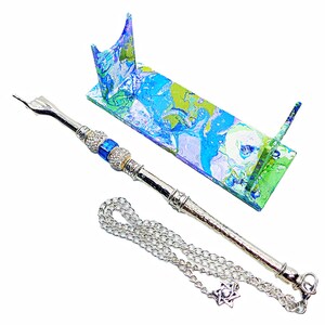 May include: A silver-toned metal Havdalah set with a Star of David pendant and a matching stand. The stand features a colorful abstract design in shades of blue, green, and white. The set includes a chain and a decorative element with blue gems.