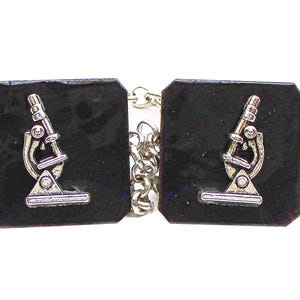 May include: A pair of black square cufflinks with a silver microscope design. The cufflinks are connected by a silver chain.