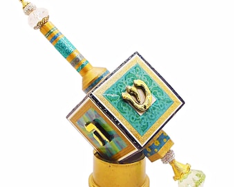 May include: A decorative dreidel with a gold base and accents. The dreidel features a square body with teal and gold detailing, and a gold letter on each side. The handle is yellow with blue and glitter stripes, and a clear, faceted top.