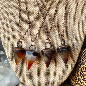 Puede incluir: Cuatro collares de cadena de cobre con colgantes triangulares. Los colgantes son una mezcla de colores marrón, blanco y rojo. Los collares se exhiben sobre una superficie de tela beige.