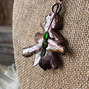 Può includere: Una collana con ciondolo a forma di foglia color rame con tre pietre preziose verdi. Il ciondolo è una rappresentazione dettagliata di una foglia di quercia, con venature visibili e una superficie strutturata. La catena della collana è anch'essa color rame.