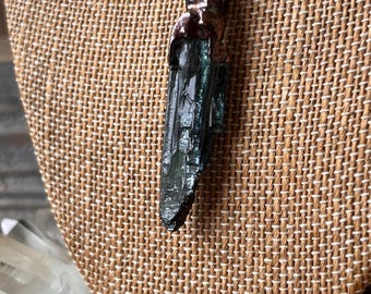 May include: A raw, dark green, elongated crystal pendant suspended from a copper-colored metal chain. The crystal features a vertical, striated texture and is secured at the top by a dark, metallic cap with an intricate, swirling design.